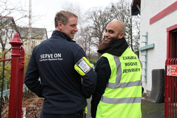 Two men, one in a dark jacket and another in a high-visibility vest, smiling and conversing outside.