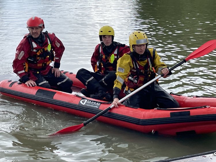 Three people in a small inflatable boat on a calm river, wearing red and yellow safety gear.