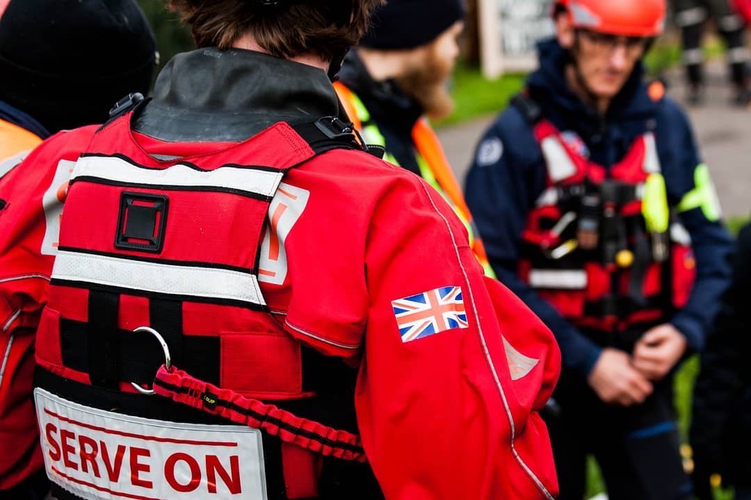 Close-up of a rescue worker’s back, showing a red jacket with a UK flag and "SERVE ON" patch.