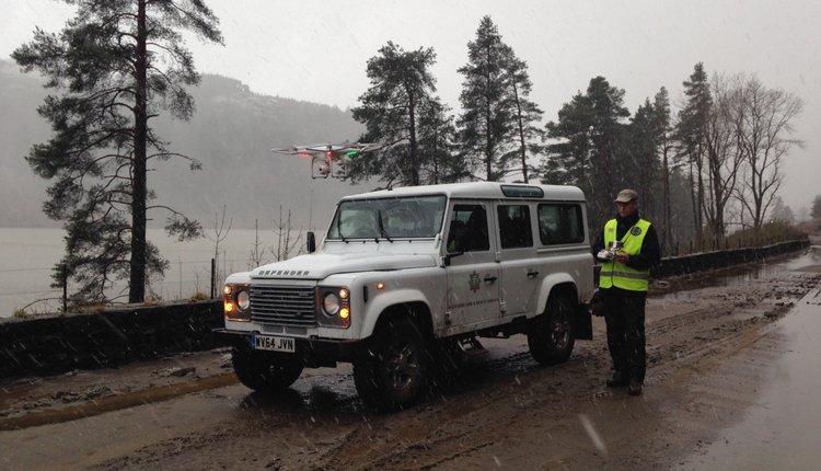 A person operating a drone beside a 4x4 vehicle in a rainy, forested area.