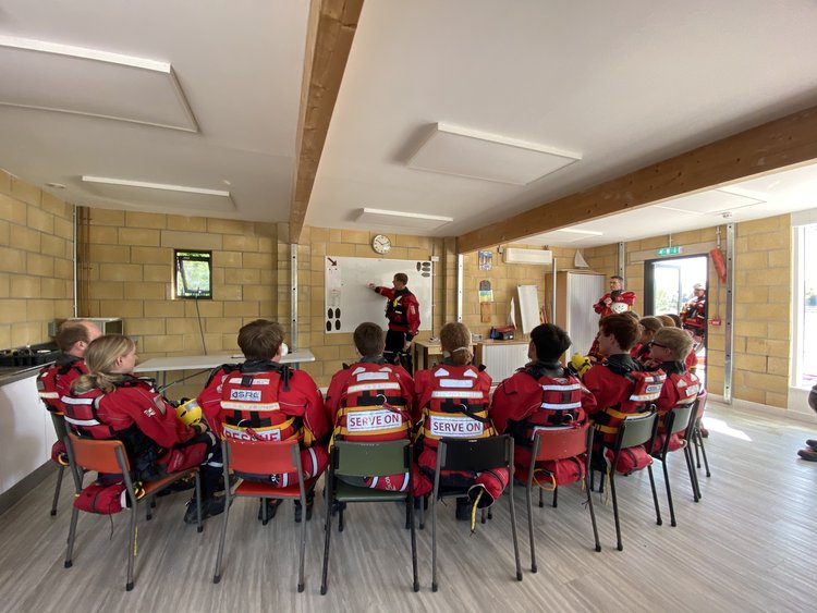Indoor training session with a group of people in red jackets seated and listening to a presenter.
