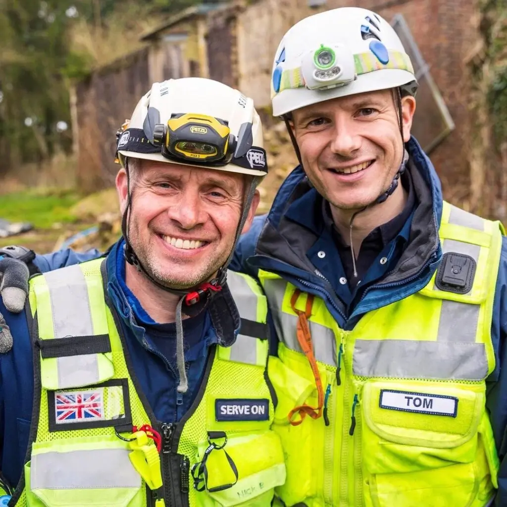 Two smiling rescue workers in high-visibility jackets with helmets and name tags.
