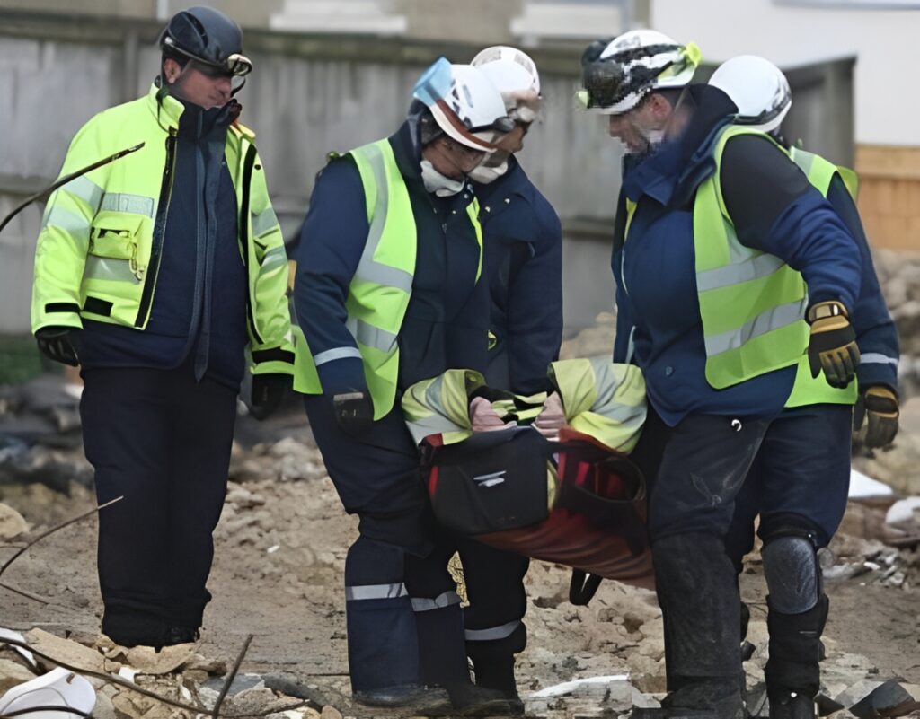 Three emergency responders carrying a stretcher with a casualty in a debris-covered area.