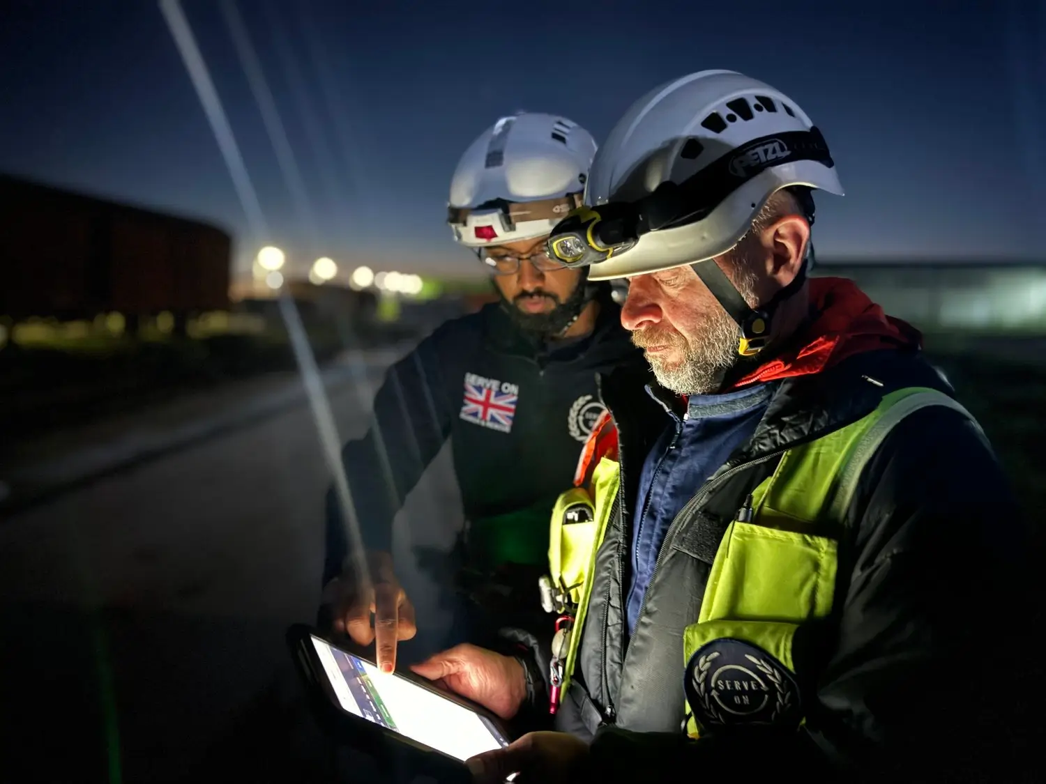 Two search and rescue members reviewing information on a tablet at night with industrial lights in the background.